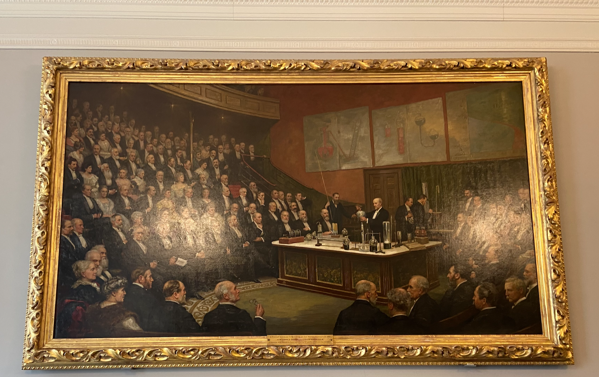 Rectangular oil painting with golden frame, lecture hall full of older, white men, red walls, three pictures above the wooden table in the centre and James Dewar behind the lecture table