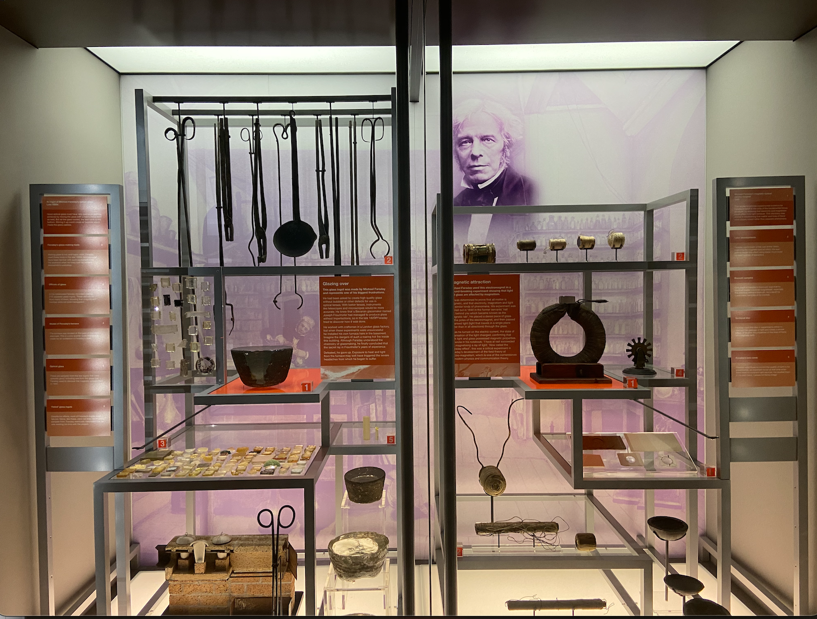 Lots of scientific equipment behind glass panes in two sections, white walls with one purple pictures of a scientist, clear flooring, 11 orange rectangular captions, two orange square captions and metal shelving