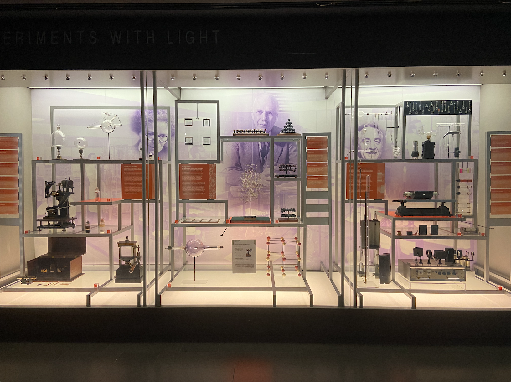 Lots of scientific equipment behind glass panes in three sections, white walls with three purple pictures of scientists, clear flooring, 16 orange rectangular captions, three orange square captions and metal shelving