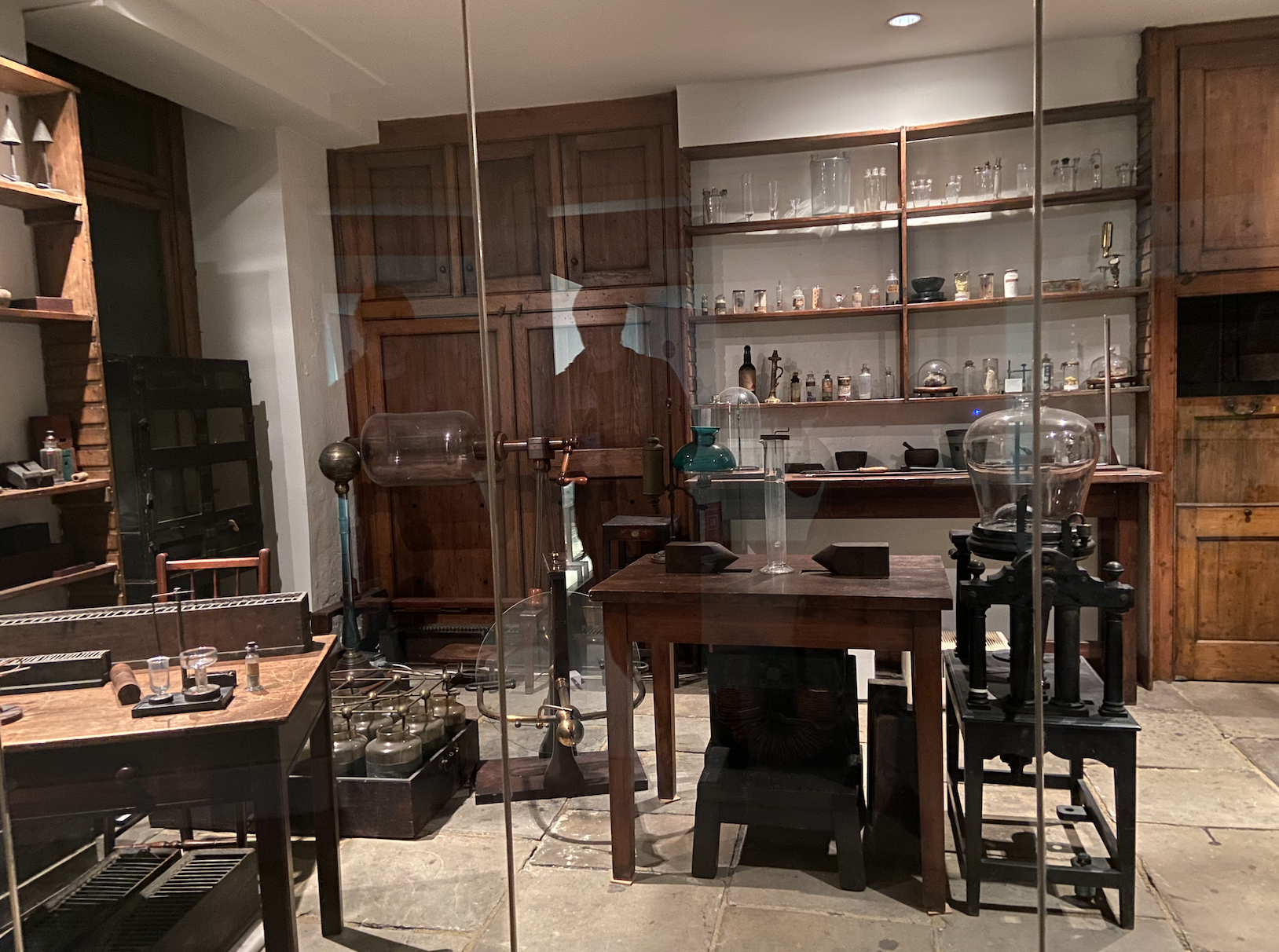 A lab with white walls behind panes of glass, grey stone flooring, two wooden tables, two wooden chairs, three bookshelves full of clear glass vials and bottles, two wooden doors, one circle light, a green lamp and scientific equipment scattered around