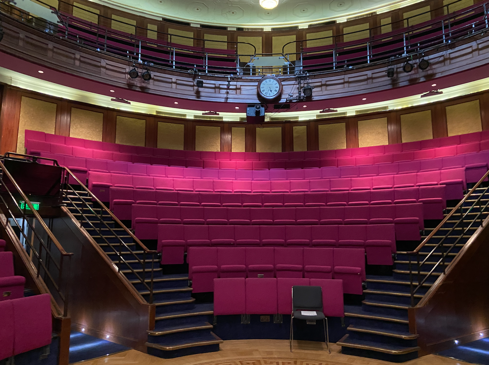A front view of a lecture hall with red seats, a clock in the middle, a balcony and 2 sets of stairs