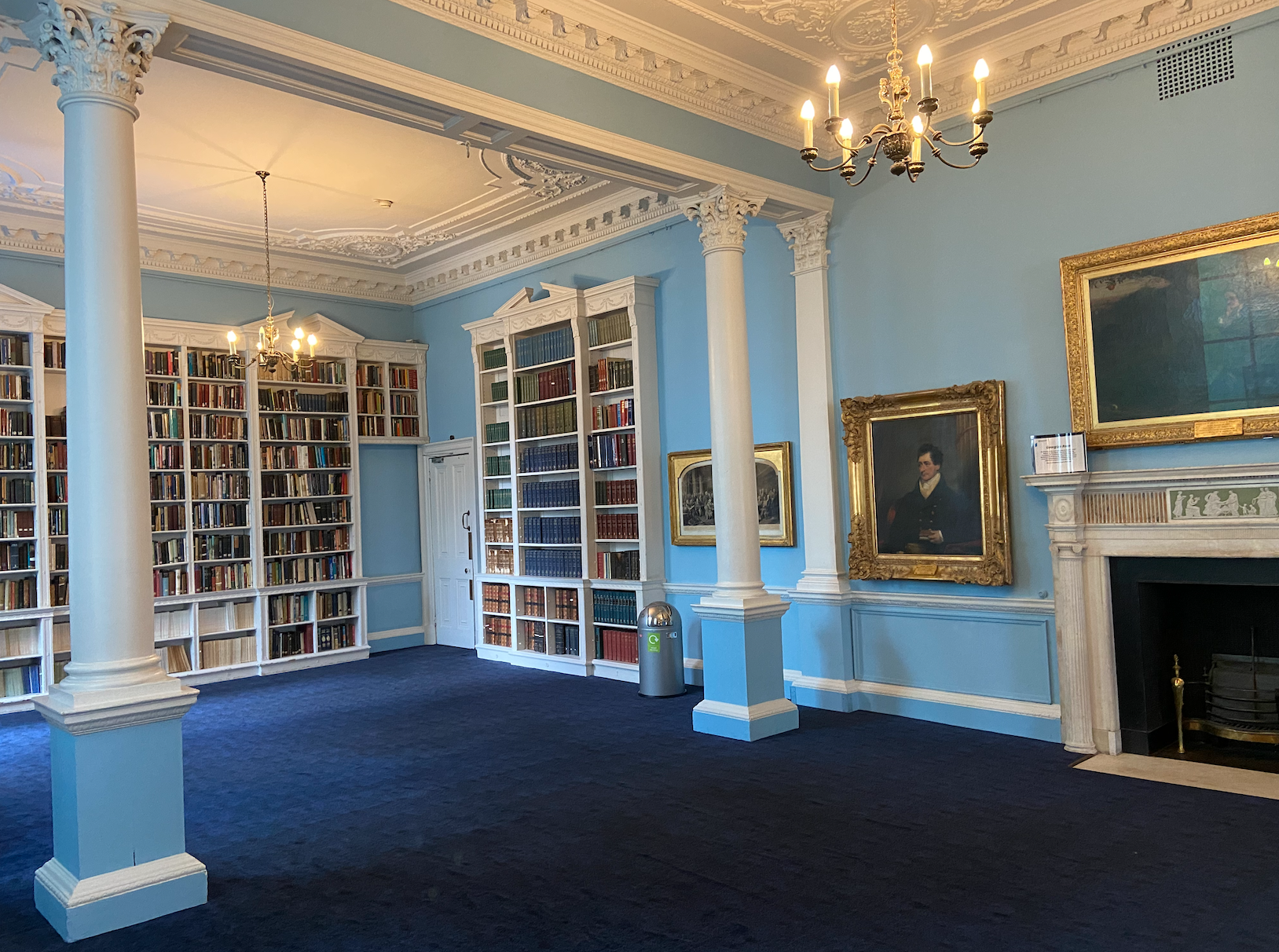 Library with light blue wallpaper, dark blue carpet flooring, three full bookcases, two white columns, a white and black fireplace, two chandeliers and three paintings on the walls