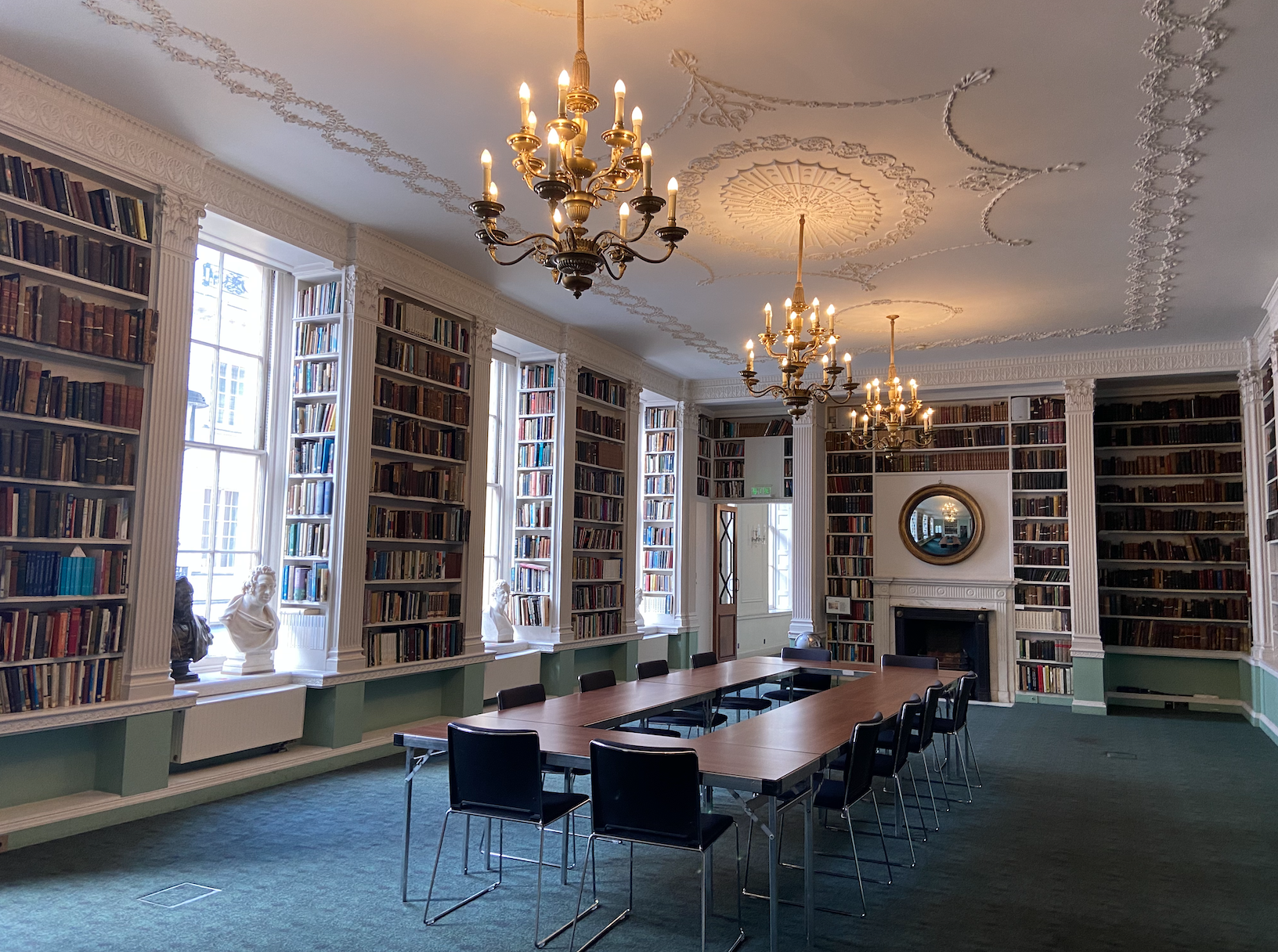 Library with white walls, blue carpet flooring, nine full bookcases, a white and black fireplace, three chandeliers, a brown rectangular table with a smaller rectangular hole in the middle, 12 blue chairs with metal legs around the table and four white busts of scientists