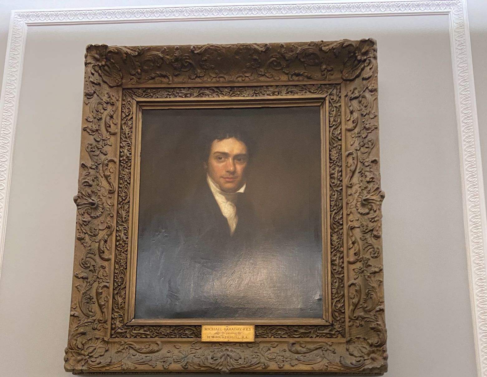 A square painting of Michael Faraday with an intricate wooden frame, standing up, white walls, white middle aged man, brown hair, black waistcoat and white cravat