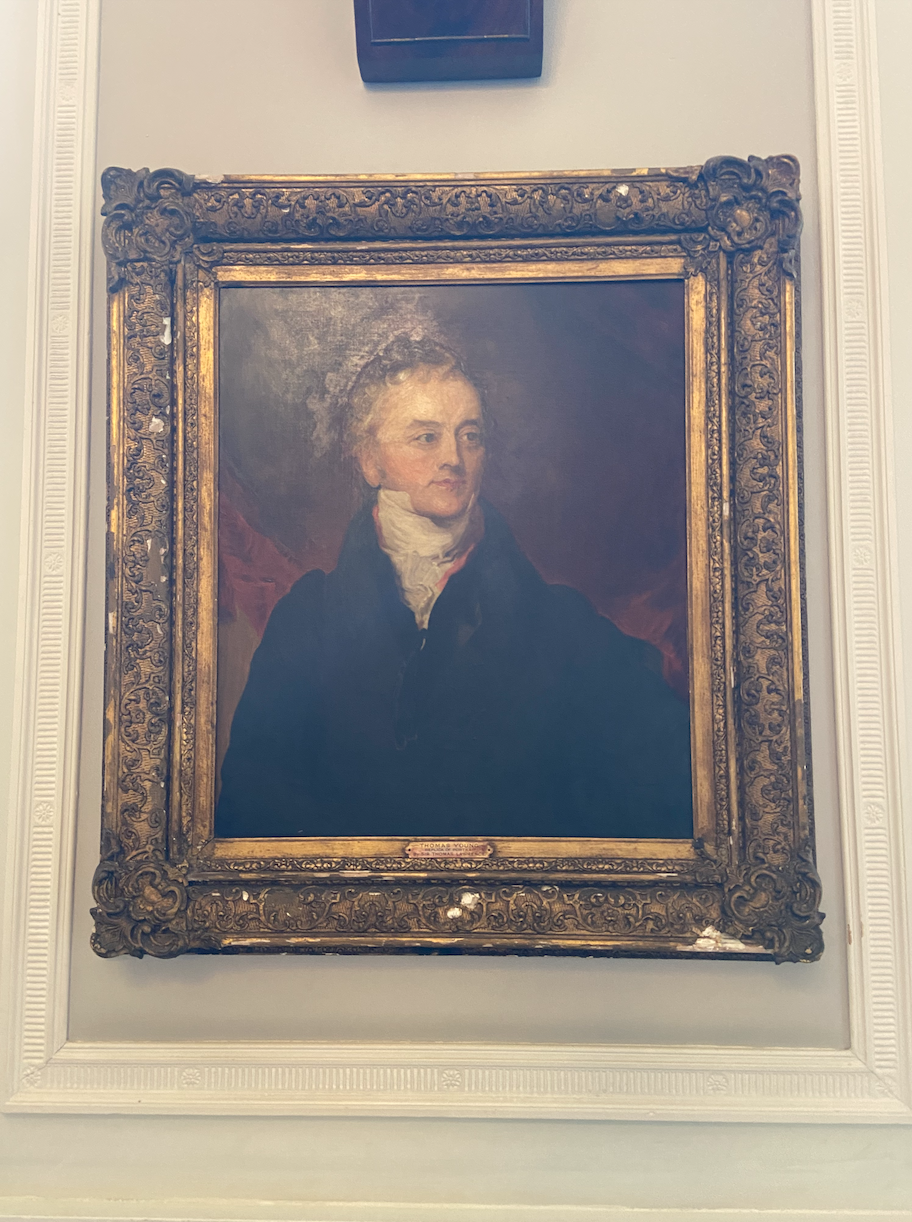 A square painting of Thomas Young with a faded golden frame, standing up, white walls, white older man, grey hair, black waistcoat and white cravat